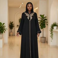 Woman wearing a black abaya with white embroidery in a well-lit room with plants.